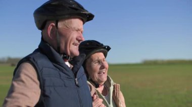 Bisikletli yaşlı bir çift şehrin dışında spor yürüyüşü yapıyor. Bir kadın bir erkeğin elini tutuyor ve onunla konuşuyor. Çift güzel güneşli yeşil çayırların arka planında yürüyor.