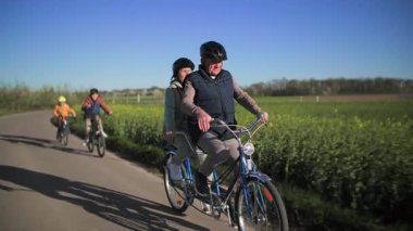 İki koltuklu bisiklete binen yaşlı bir çift ve torunları tarlalar arasında bir kır yolu boyunca bisiklet sürüyor, ve ılık bahar güneşinin ışınlarında yeşil çayırlar 