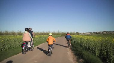 İki koltuklu bisiklete binen yaşlı bir çift ve torunları sıcak bahar güneşinin ışınlarında yeşil çayırlar, tarlalar ve meyve ağaçları arasında bir kır yolu boyunca bisiklet sürüyorlar. Çekim görünümü