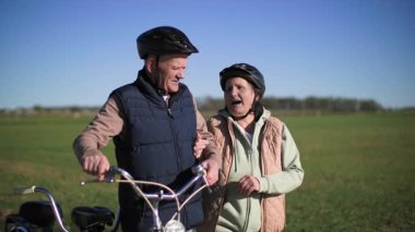 Bisikletli yaşlı bir çift şehrin dışında spor yürüyüşü yapıyor, yaşlı bir adam bisiklet sürerken sarılıyor yeşil çayır arka planında güneşli bahar havasında