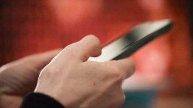 Kadın eli cep telefonunu tutar ve parmaklarıyla yazar. Renk şeması, siyah ve beyaz aksanlı, kahverengi ve turuncu gibi sıcak tonları içerir. Genel sahne bir şey ifade ediyor.
