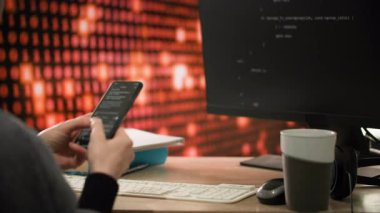Kadın bir programcı bir telefon tutar ve üzerine bir program kodu yazar, bu da monitör ekranında görüntülenir. Masanın üzerinde bir fincan kahve, bir fare ve kulaklıklar var. Arka planda bir bilgisayar var.
