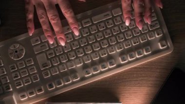 Güzel manikürlü, bakımlı kadın elleri karanlık ortamda beyaz bir klavyeye yazı yazıyor. Ofis masasında klavyenin yanında içinde kalem ve telefon olan bir not defteri var. Üst Manzara. Çok yakın.