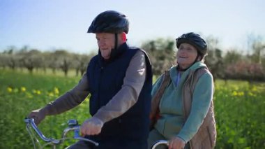 Yaşlı bir çift, karı koca, kask takıyor, yeşil tarlaların arka planında, güneşli baharda, iki koltuklu bisiklete biniyorlar. Gezi hakkında hevesle tartışıyorlar.