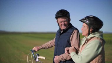 Şehrin dışında sportif bir yürüyüşte, bisikletli yaşlı bir çift yeşil çayır ve kır çiçekleriyle birbirlerine sarılarak yürüyor. Orta uzunlukta..