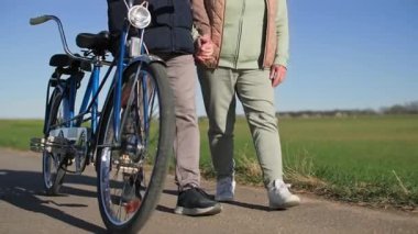 Bisikletli yaşlı bir çift şehir dışında spor gezisi yapıyor. Aynı anda hareket eder ve güneşli bahar havasında yeşil çayırların arka planında el ele tutuşurlar. Aşağıdan görüntüle
