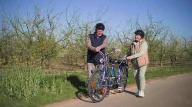 Bisiklet durağında mola verdikten sonra, yaşlı bir çift bisikletlerini kırsal bir yola yuvarlayarak bisiklet sürmeye hazırlandılar. Başlangıç prosedürünü tartışırken. Orta uzunlukta..