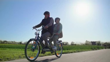 İki koltuklu bisiklet süren yaşlı bir çift yeşil tarlaların arasında şehir dışına çıkıyor. Arka planda, güneş ışığı altında alçak bir yerde çekim. Düşük ihtimal. Yavaş çekim