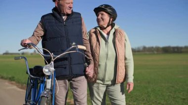 Bisikletli yaşlı bir çift şehir dışında spor gezisi yapıyor. Aynı anda hareket eder ve güneşli bahar havasında yeşil çayırların arka planında el ele tutuşurlar. Kamera hareket ediyor.