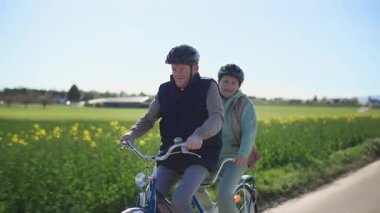 Aktif bir yaşam tarzı sürdüren yaşlı bir çift iki koltuklu bisiklette spor yapıyor. Onlar sıcak bahar güneşli havada güzel bir kırsal alanda bir yol boyunca at sürüyorlar. Bir sürü...