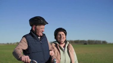 Bisikletli yaşlı çift şehrin dışında spor yürüyüşü yapıyor. Yürüyen, yaşlı bir adam konuşuyor ve güneşli bahar havasında yeşil çayır arka planında kadına sarılıyor. Orta boy.. 