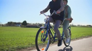 Miğfer takan yaşlı bir çift, karı koca, sıcak bahar güneşli havalarda, kırsal bir zeminde iki koltuklu bisiklete biniyor. Bir sürü izlenim ediniyorum.