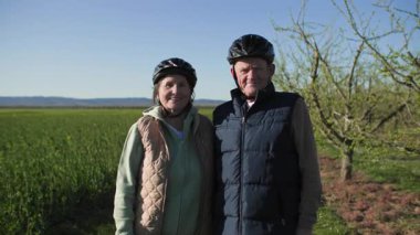 Emekli çift, kırsal kesimdeki çiçek tarlaları ve meyve ağaçlarına karşı birlikte poz veriyor. Parlak güneşli bir günde ve ılık bahar havasında