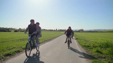 İki koltuklu bisiklet süren yaşlı bir çift, torunlarıyla birlikte yeşil tarlaların arasında şehir dışında geziyorlar. Bisikletli torunlar birlikte gezmeye devam etmek için büyükbabalarına yetişirler.