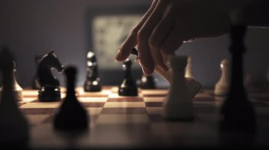 Bir adam zarif bir şekilde beyaz bir veziri satranç tahtasının ortasına yerleştirir ve zarafetle onu işaret eder. Arka planda, bir saatin tik tak ettiği görülebilir ve aciliyet duygusuna eklenebilir.