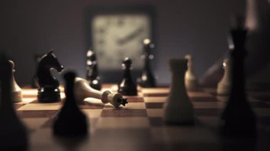 Beyaz bir kral satranç taşları arasında bir satranç tahtasında sallanıyor. Tıklayan bir saat arka planda görülebilir, stratejik bir oyunun hissini güçlendirir. Arka planda başka satranç taşları var.