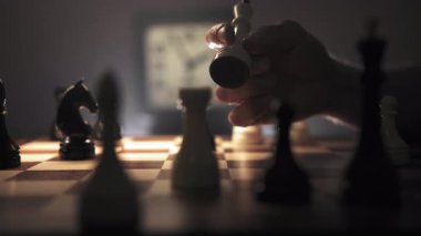 Bir adam elinde vezir taşını satranç tahtasının tepesinde çeviriyor. Arka planda, duvarda beyaz bir saat asılı. Fotoğraf bir konsantrasyon ve stratejik düşünme hissi veriyor.