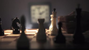 Adam satranç oynamaya derinden odaklanmış. Önündeki satranç tahtası ve hareket eden el satranç taşını hareket ettirmek üzere. Arka planda tıklayan bir saat görülebilir, eklenerek