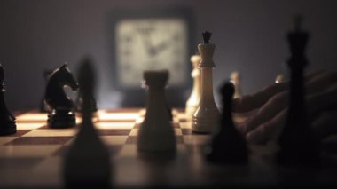 Satranç tahtasının ve üzerindeki elin satranç taşını hareket ettirmeye hazırlandığı gibi adam satranç oynamaya derinden odaklanmış durumda. Arka planda tik tak yapan bir saat görülebilir,