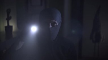 Siyah maskeli ve siyah elbiseli bir adam loş bir odada. Yüzü kısmen gizlenmiş, ona gizemli bir görünüm veriyor. Elinde bir el feneri tutuyor, araştırmak için kullanıyor.