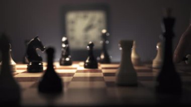 Erkek eli, beyaz bir veziri satranç tahtasının ortasına koyar ve zarifçe parmağını ona doğrultur. Arka planda, bir saatin tik tak ettiği görülebilir, stratejik oyunun aciliyetini artırır