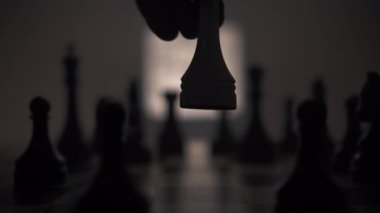 Bir satranç tahtasına çok sayıda siyah taşla yakın plan, ortasında bir insan eli sinsice beyaz bir papaz taşını yerleştiriyor. Satranç tahtasının arkasında parlak siyah-beyaz bir turnuva var.