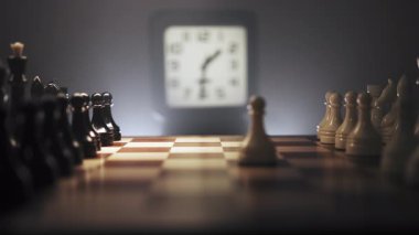 Satranç tahtasının yanında saati gösteren bir saat var. Sahne sıcak ışıkla aydınlatılıyor, sıcak ve yoğun bir atmosfer yaratıyor. Çekim, stratejik düşünme ve konsantrasyon anını gösteriyor.