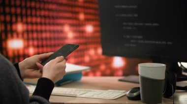 Zaman turları. Bir programcı bir telefon tutar ve üzerine bir program kodu yazar, bu da monitör ekranında görüntülenir. Masanın üzerinde bir fincan kahve, bir fare ve kulaklıklar var. Arka planda bir...