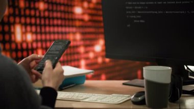 Bir programcı bir telefon tutar ve üzerine bir program kodu yazar, bu da monitör ekranında görüntülenir. Masanın üzerinde bir fincan kahve, bir fare ve kulaklıklar var. Arka planda bir bilgisayar monitörü var.