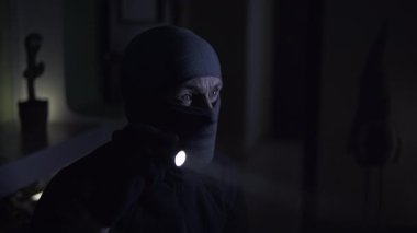 Siyah maskeli ve siyah elbiseli bir adam loş bir odada duruyor. Yüzü kısmen gizlenmiş, ona gizemli bir görünüm veriyor. Elinde bir el feneri var, onu arama yapmak için kullanıyor.