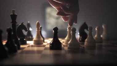 Satranç tahtası çeşitli taşlarla kurulur ve insan eli tahtada taşları hareket ettirirken görülür. Arka planda, duvarda beyaz bir saat var. İğne bir konsantrasyon hissi yayıyor.