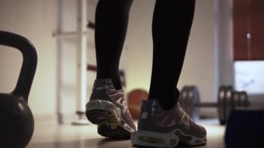 Bir adam spor salonunda yerde ısınıyor ve antrenman için hazırlanıyor. Adam spor ayakkabı giyiyor, tabanları çerçevede açıkça görülebiliyor. Adamın ayaklarının yanında bir çaydanlık çanı var.