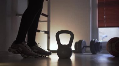 Spor ayakkabılarıyla spor salonunda sporcu ayakta, ısınıyor ve vücudunu rahatlatıyor. Ön planda, önünde yerde bir kettlebell var. Spor salonundaymış ve spor yapıyormuş.