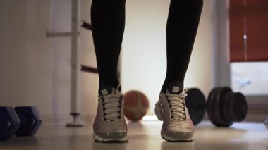 Spor ayakkabılı bir sporcu, atlayıp vücudunu rahatlatarak ısınıyor. Ön planda ve arka planda yanında dambıllar var. Bir fitness merkezinde ve antrenman yapıyor. Sporcular.