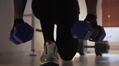 Sporcu diz çöker ve dambılları yere yerleştirir, sonra onları tekrar kaldırır. Arka planda onun yanında yerde büyük bir dambıl var. Spor salonunda ve antrenman yapıyor.