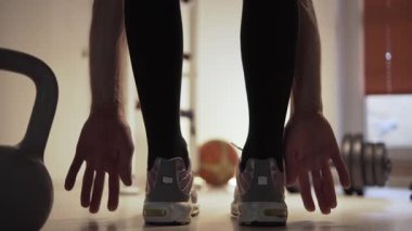 Spor ayakkabılı bir sporcu, vücudunu esnetiyor ve gevşetiyor. Önünde bir kettlebell, arkasında da yerde bir dambıl var. Spor salonundaydı ve...