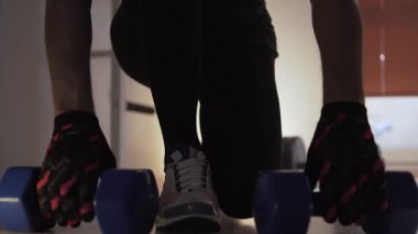 Sporcu diz çöker, dambılları eline alır ve öne doğru eğilir. Arka planda onun yanında yerde bir dambıl var. Bir fitness merkezinde ve antrenman yapıyor. Sporcu hareketleri