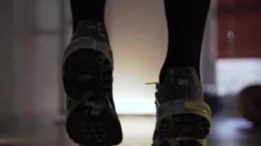 Spor salonunda spor ayakkabı giyen bir adam antrenmandan önce koşarak ısınıyor. Spor yaptığı bir fitness merkezinde kalıyor. Dambıl ve basketbol topu arka planda görünür.