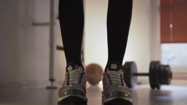 Sporcu, dambılları eline alır ve öne doğru eğilir. Arka planda onun yanında yerde bir dambıl var. Bir fitness merkezinde ve antrenman yapıyor. Atletizm hareketleri