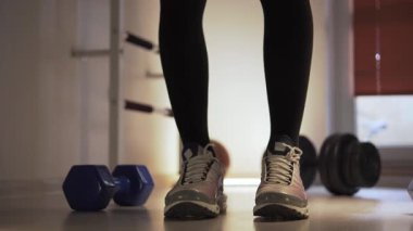 Spor ayakkabılarını giymiş, ayakta duran, esneyen ve vücudunu rahatlatan bir sporcu. Ayaklarını ısıtıyor, bacaklarıyla hareket ediyor. Ön planda yanında duran dambıllar var.