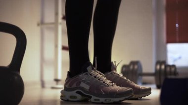 Bir adam spor salonunda yerde ısınıyor ve antrenman için hazırlanıyor. Adam spor ayakkabı giyiyor, tabanları çerçevede açıkça görülebiliyor. Adamın ayaklarının yanında bir çaydanlık çanı var.