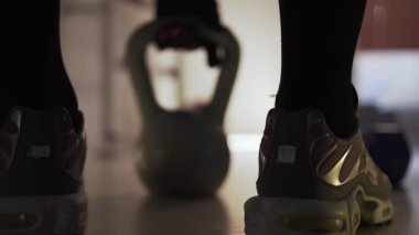 Spor salonunda, spor ayakkabılı bir adam bir ağırlık alıp kaldırır. Spor yaptığı bir fitness merkezindeymiş. Arka planda birkaç dambıl var. Arkadan insan bacakları görünüyor.