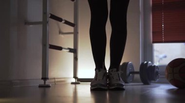 Spor ayakkabılı bir sporcu, bir su ısıtıcıyla atlama yapıyor. Antrenman yaptığı bir fitness merkezinde. Arka planda bir dambıl ve bir basketbol topu var. Erkek bacaklarının görüntüsü...