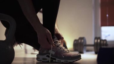 Atletik yapılı bir adam spor salonunda dizlerinin üzerinde duruyor ve spor ayakkabılarının bağcıklarını bağlıyor. Adam bağcıkları bağlamaya odaklanmış, ki bu da olayın detaylarını ve gelişigüzel...