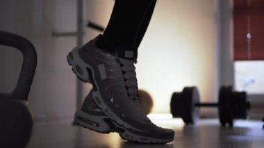 Spor ayakkabılı bir adam kendini antrenmana hazırlamak için çeşitli ısınma hareketleri yapar. Önünde bir kettlebell, arkasında da yerde bir dambıl var. Formda.