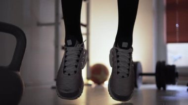 Spor salonunda ısınan ve antrenmana hazırlanan bir adam. Koşarak ve zıplayarak ısınıyor. Adam spor ayakkabı giyiyor, tabanları çerçevede açıkça görülebiliyor. Şunun yanına.