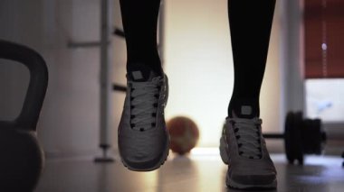 Spor salonunda ısınan ve antrenmana hazırlanan bir adam. Koşarak ve zıplayarak ısınıyor. Adam spor ayakkabı giyiyor, tabanları çerçevede açıkça görülebiliyor. Şunun yanına.