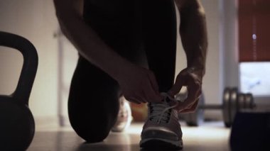 Atletik yapılı bir adam spor salonunda dizlerinin üzerinde duruyor ve spor ayakkabılarının bağcıklarını bağlıyor. Adam bağcıkları bağlamaya odaklanmış, ki bu da olayın detaylarını ve gelişigüzel...