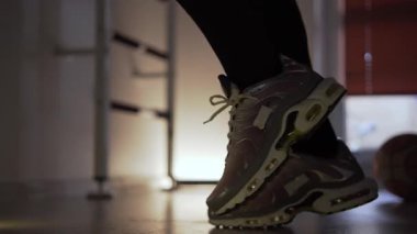 Spor salonunda spor ayakkabı giyen bir adam antrenmandan önce koşarak ısınıyor. Spor yaptığı bir fitness merkezinde kalıyor. Dambıl ve basketbol topu arka planda görünür.