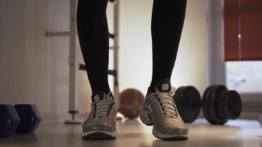 Spor ayakkabılarını giymiş, ayakta duran, esneyen ve vücudunu rahatlatan bir sporcu. Ayaklarını ısıtıyor, bacaklarıyla hareket ediyor. Ön planda yanında duran dambıllar var.